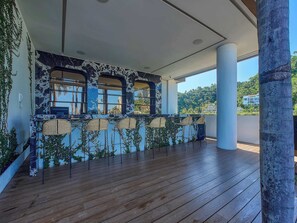 Ein überdachter Außenbereich mit Holzboden, eine Bar mit Korbstühlen und Blick auf einen Hügel.