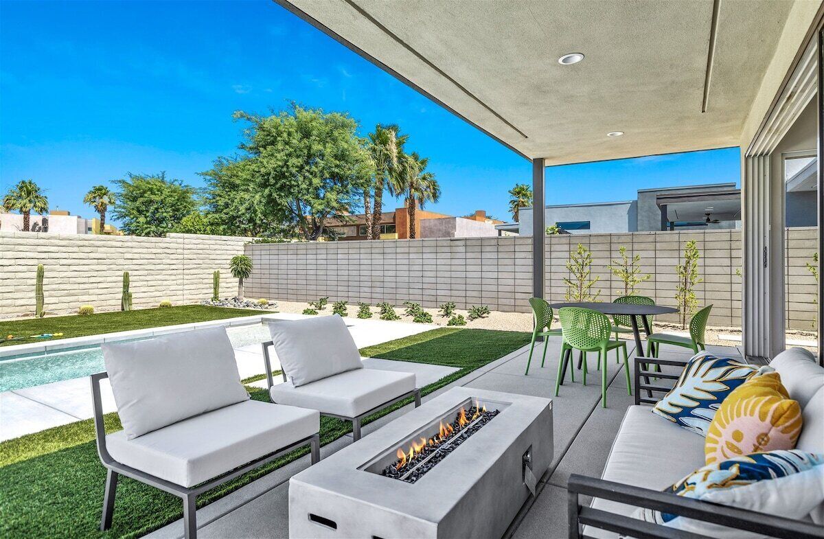 A modern outdoor patio with a pool, white lounge chairs, a fire pit, and a dining area with green chairs.