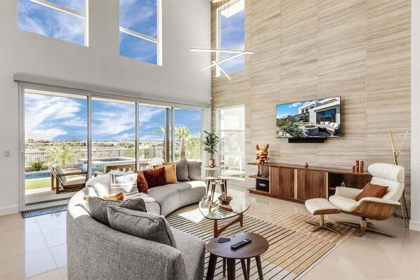 A spacious living room with large windows, a modern sofa, a coffee table, and a TV mounted on the wall.
