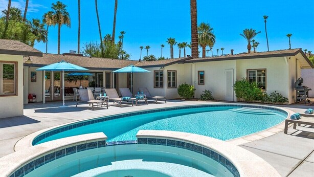 A swimming pool with lounge chairs and umbrellas, surrounded by palm trees and a house.