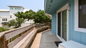 Une terrasse en bois avec une rambarde, un bâtiment bleu clair avec une fenêtre, et un arbre en arrière-plan.