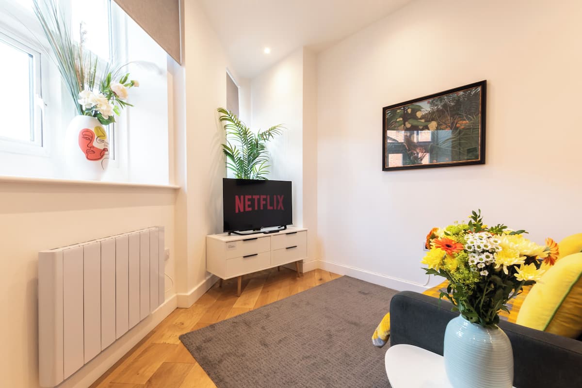 A modern living room with a white radiator, a grey rug, a sofa with yellow cushions, a blue vase with flowers, a white cabinet with a Netflix sign, and a framed picture on the wall.