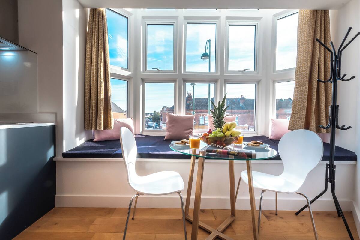 A dining area with a glass table, two white chairs, a fruit bowl, and a view through large windows.