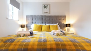 A hotel room with a large bed, a grey tufted headboard, yellow and grey checkered bedspread, two bedside tables with lamps, and a framed picture of a palm tree on the wall. - Smart Apartments - King's House (Southampton)