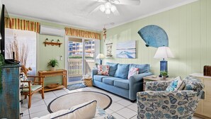 Un salon avec un canapé bleu, des fauteuils à motifs, un ventilateur de plafond et une vue sur le balcon extérieur.