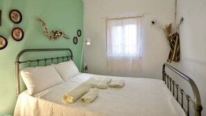 Ein Schlafzimmer mit Bett, Deckenventilator, Fenster mit durchsichtigen Vorhängen und Wanddekorationen, darunter gerahmte Bilder und getrocknete Blumen.