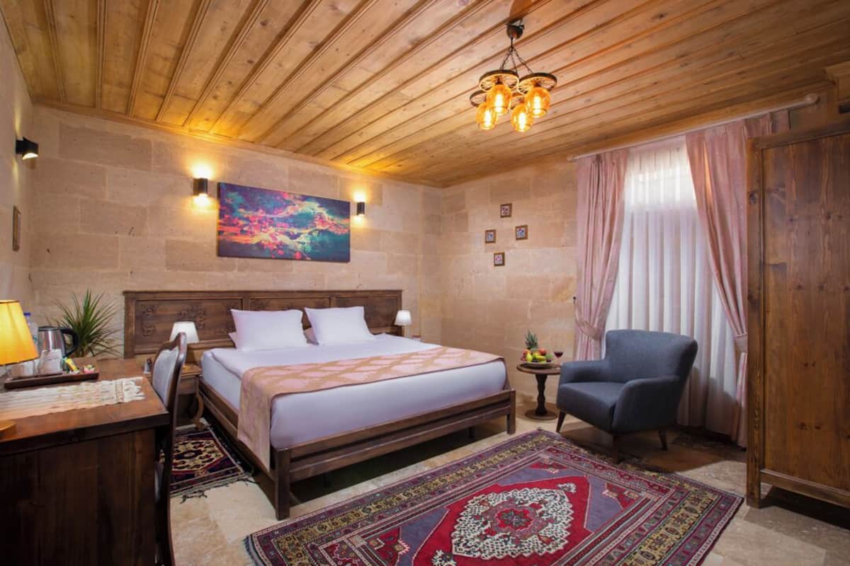 A hotel room with a wooden ceiling, a large bed with a patterned bedspread, a desk with a lamp, a chair, a small table with fruit, and a colorful painting on the wall.