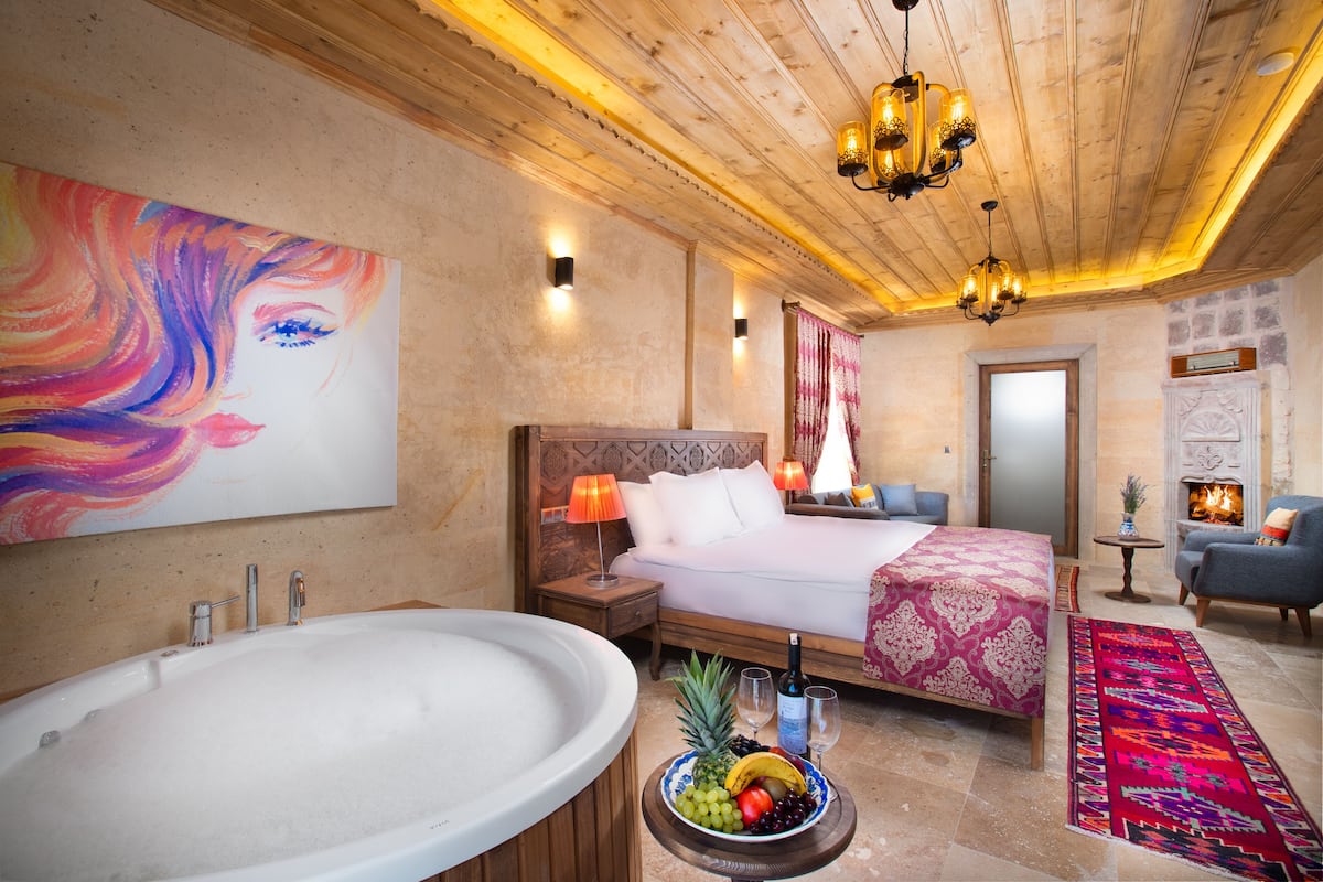 A hotel room with a large bathtub, a bed with a pink bedspread, a fruit bowl on the floor, and a fireplace.