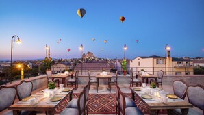 A rooftop dining area with tables set for two, overlooking a cityscape and hot air balloons in the sky. - Cappadocia Pyramid Stone House (Nevsehir)
