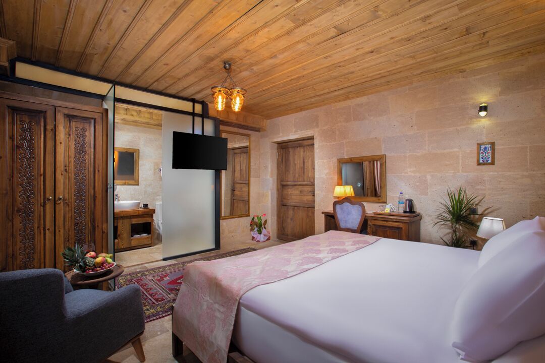 A hotel room with a wooden ceiling, a bed with a pink bedspread, a desk with a lamp, a small table with fruit, and a potted plant.
