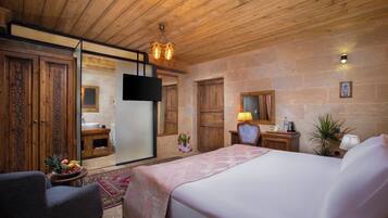 A hotel room with a wooden ceiling, a bed with a pink bedspread, a desk with a lamp, a small table with fruit, and a potted plant.