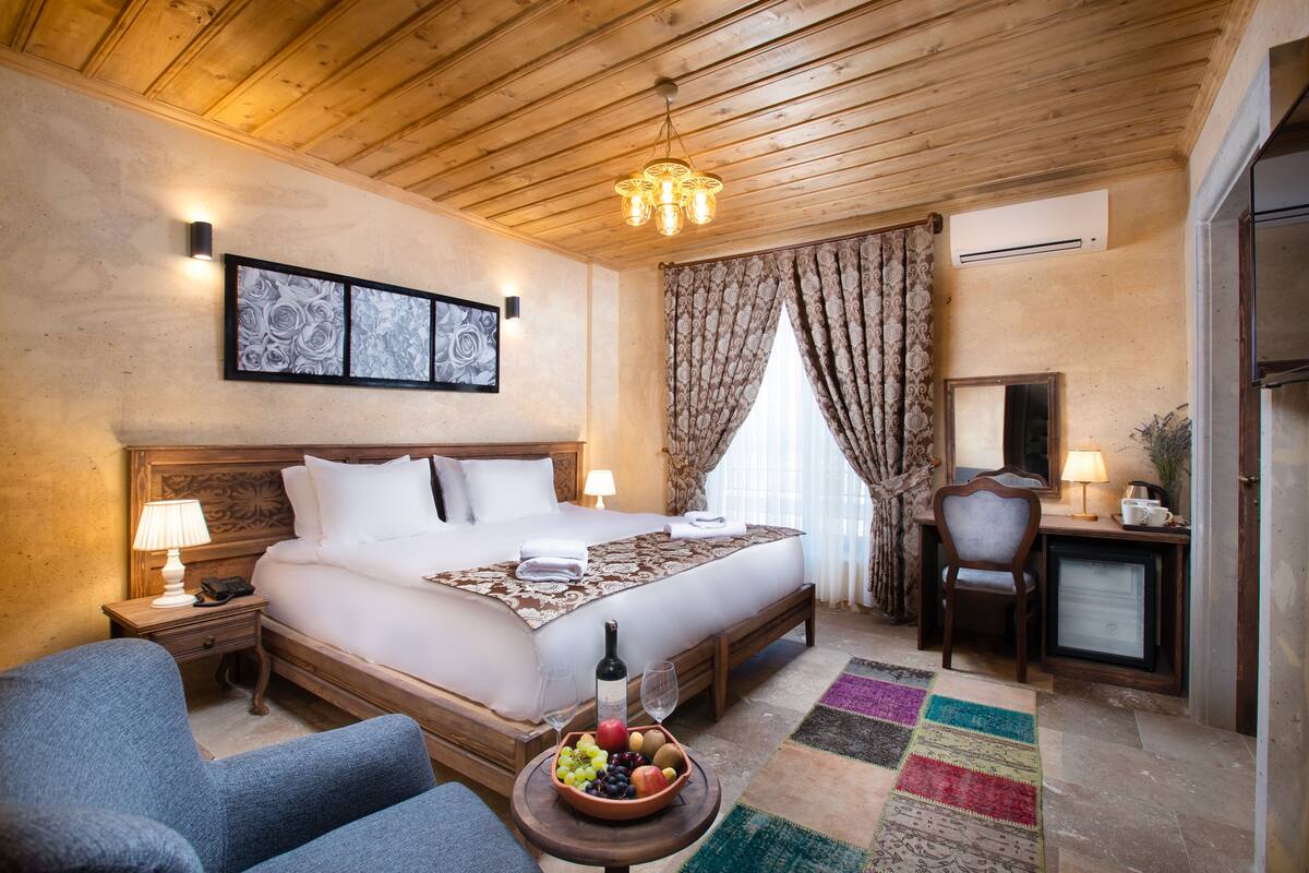 A hotel room with a wooden ceiling, a bed with white linens, a desk with a lamp, a chair, and a fruit bowl on the floor.