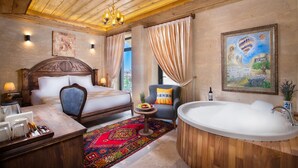 A hotel room with a wooden ceiling, a large bed, a sitting area with a chair, a bathtub, and a dining table with a fruit bowl. - Cappadocia Pyramid Stone House (Nevsehir)