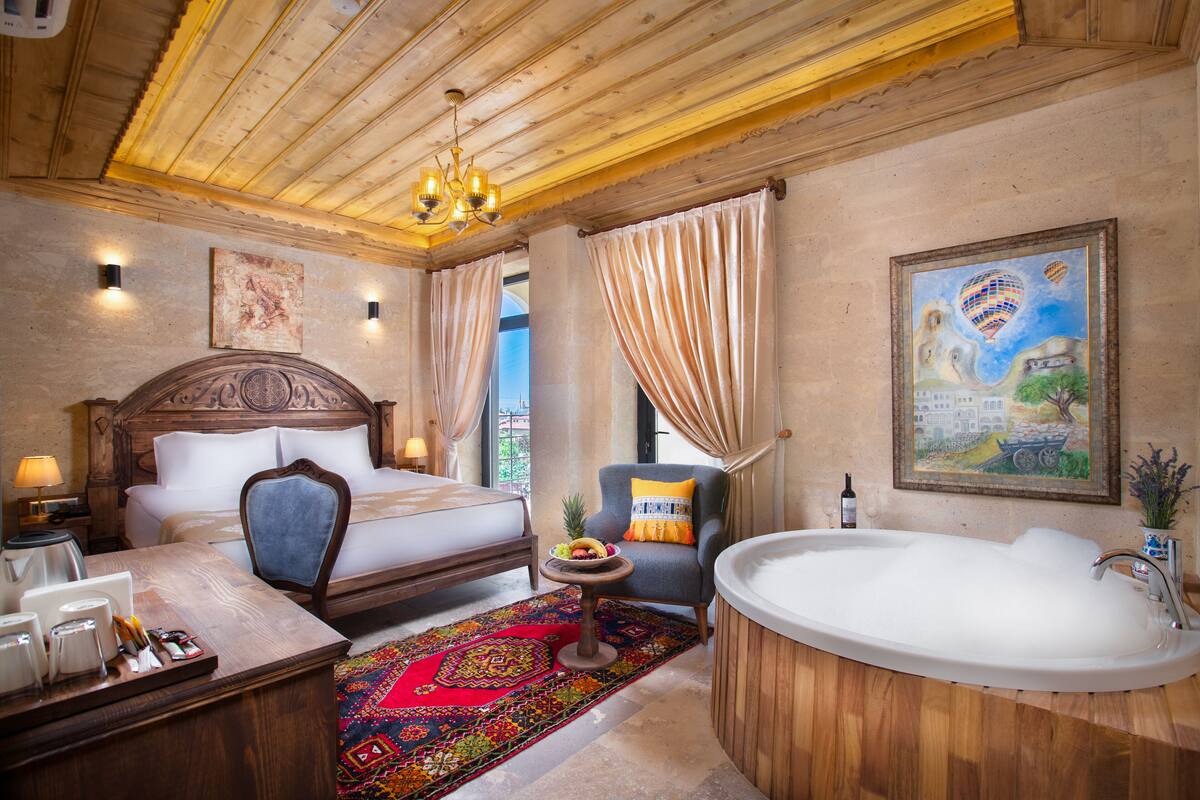 A hotel room with a wooden ceiling, a large bed, a sitting area with a chair, a bathtub, and a dining table with a fruit bowl.