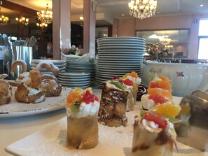 A dessert buffet with various pastries and desserts, including cream puffs and fruit-topped cakes, in a well-lit dining area with chandeliers. - Hotel Eden  (Grado)