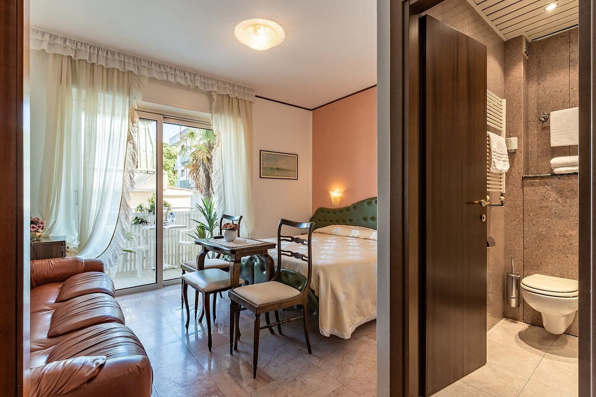 A hotel room with a bed, a dining area with a table and chairs, a sofa, and a bathroom with a toilet and towel rack.