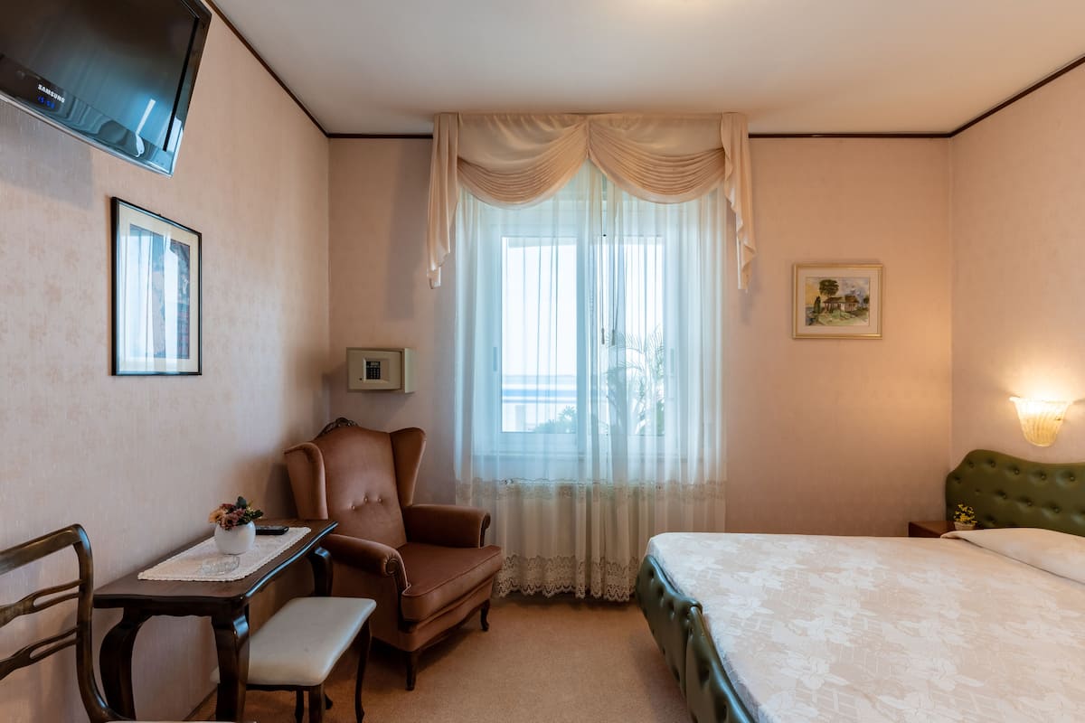 A hotel room with a bed, a desk with a chair, a television mounted on the wall, and a window with sheer curtains.