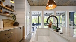 Eine moderne Küche mit Holzschränken, einer weißen Arbeitsplatte und einem großen Spülbecken. Regale mit Dekorationselementen und eine hängende Leuchte sind ebenfalls vorhanden.