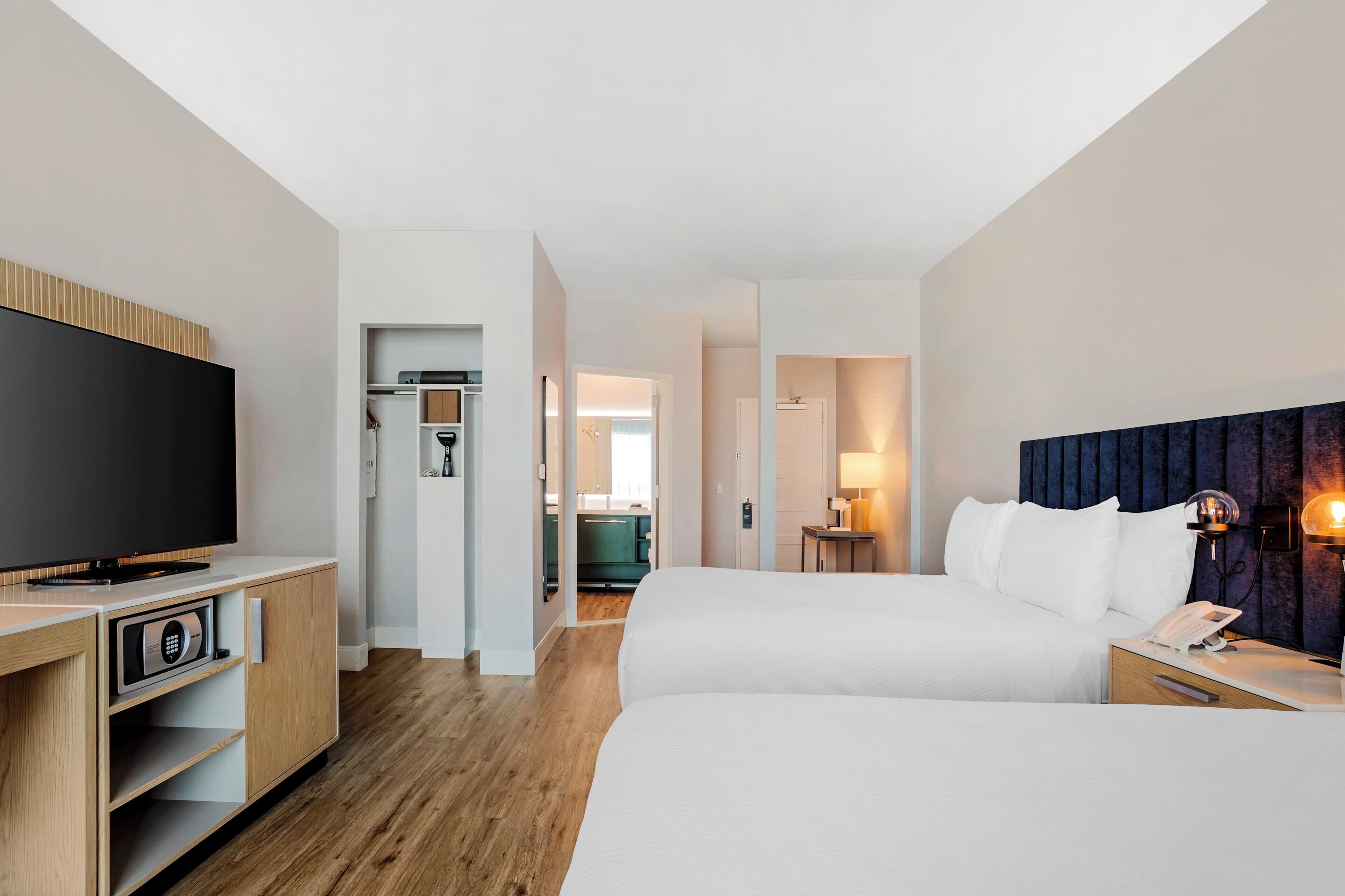 A hotel room with a flat-screen TV, a wooden cabinet, a bed with white bedding, a bedside table with a lamp, and a dark headboard.