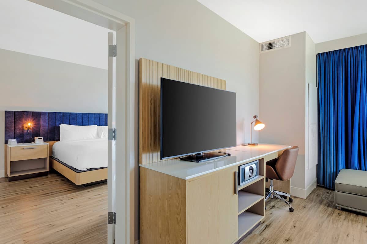 A hotel room with a bed, a desk with a TV, a chair, and a desk lamp.