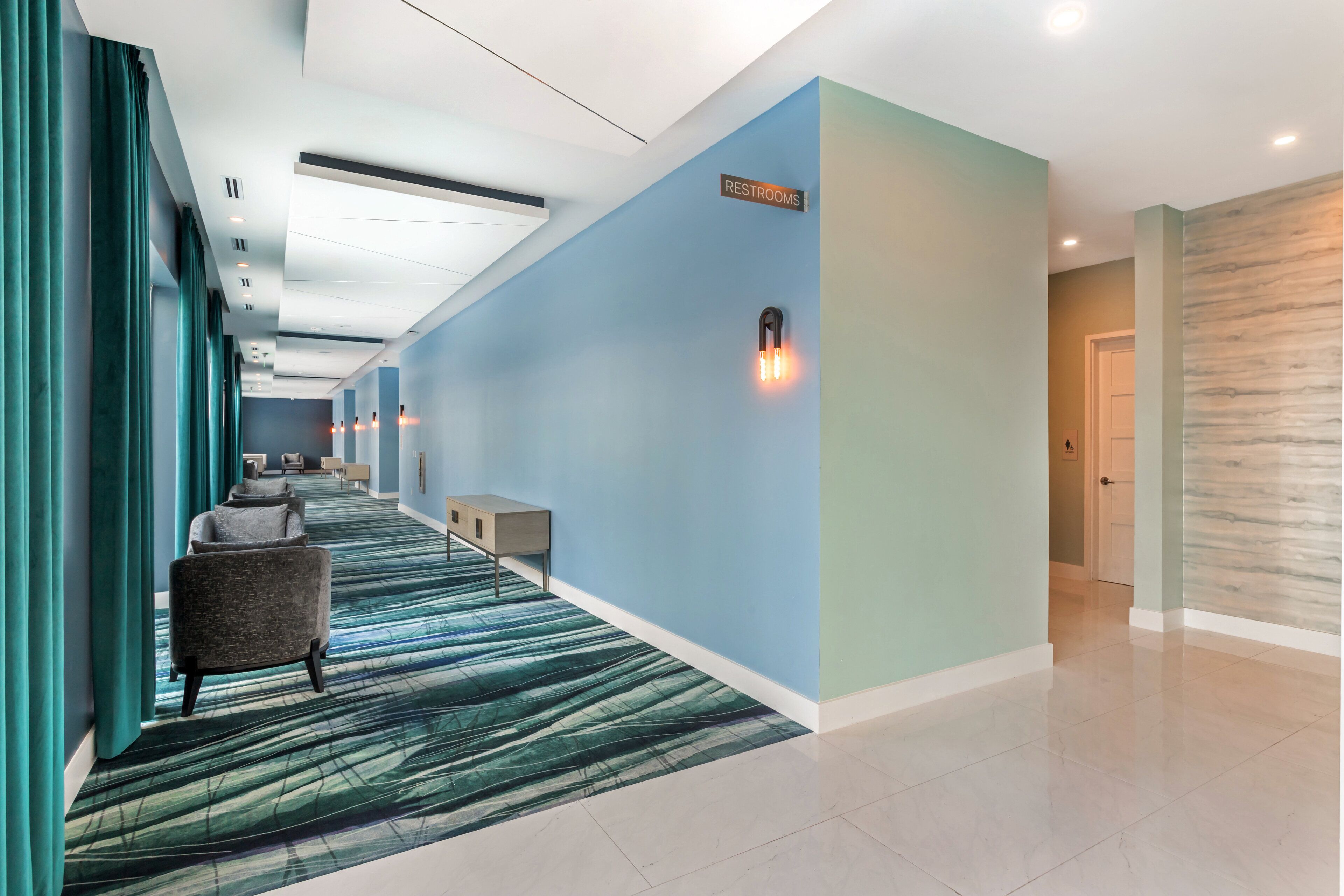 A hotel corridor with a blue wall, a sign indicating restrooms, a patterned carpet, and a bench.
