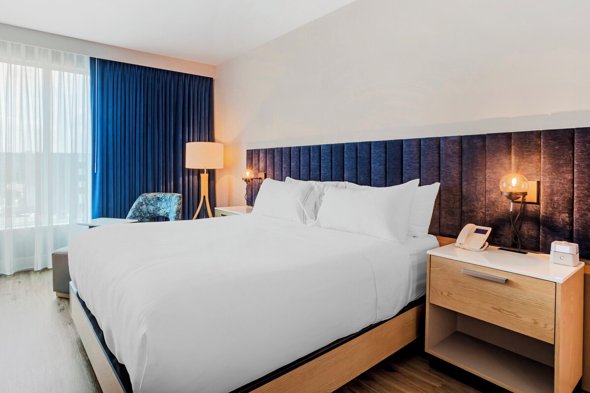 Une chambre d’hôtel avec un grand lit, une table de chevet avec une lampe, une chaise et une vue sur la ville par la fenêtre.
