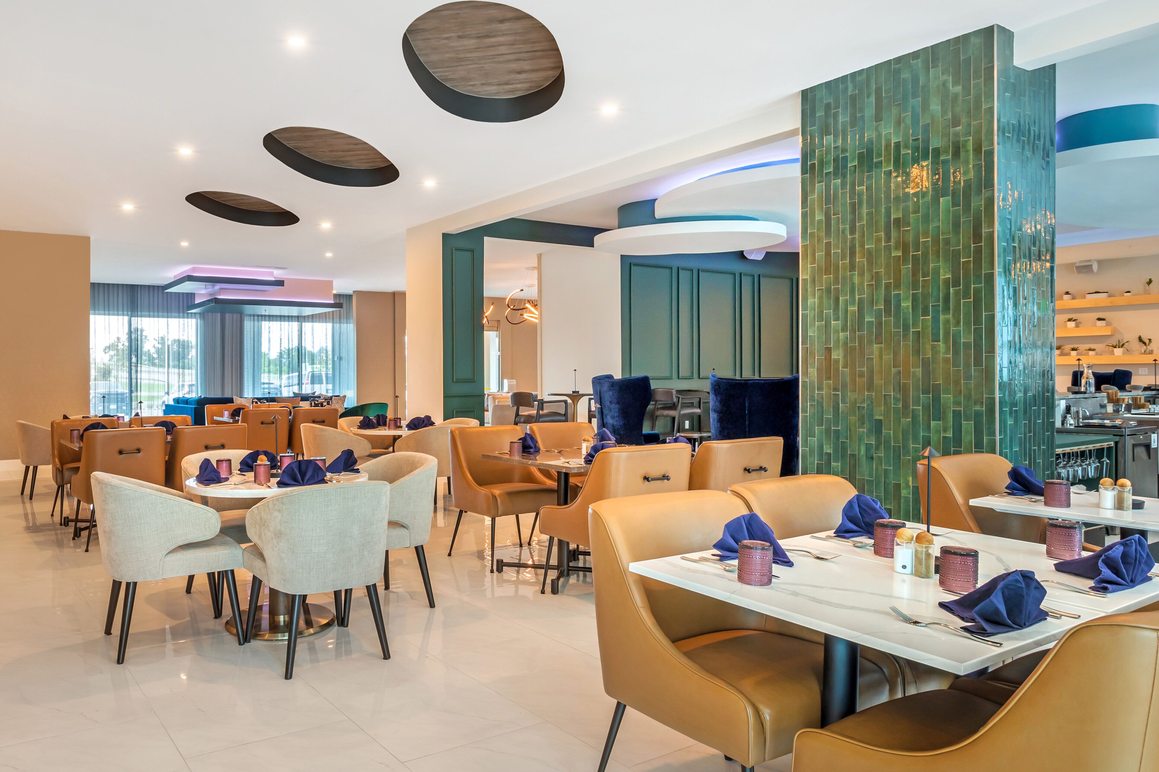 A modern dining area with round tables and chairs, a kitchen in the background, and a green textured wall.