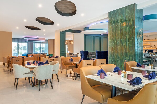A modern dining area with round tables and chairs, a kitchen in the background, and a green textured wall. - EVEN Hotel Waco - University Area by IHG (Waco)