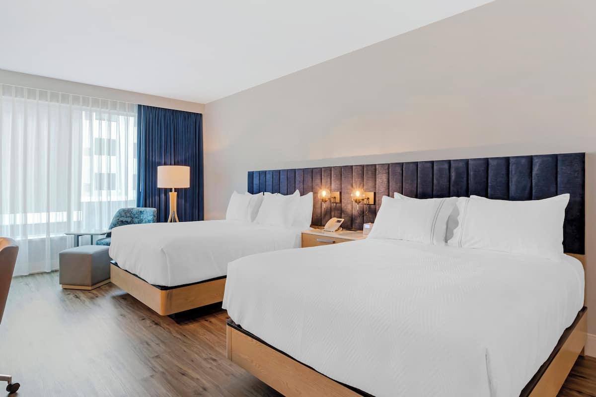 A hotel room with two beds, a desk, a chair, and a lamp.