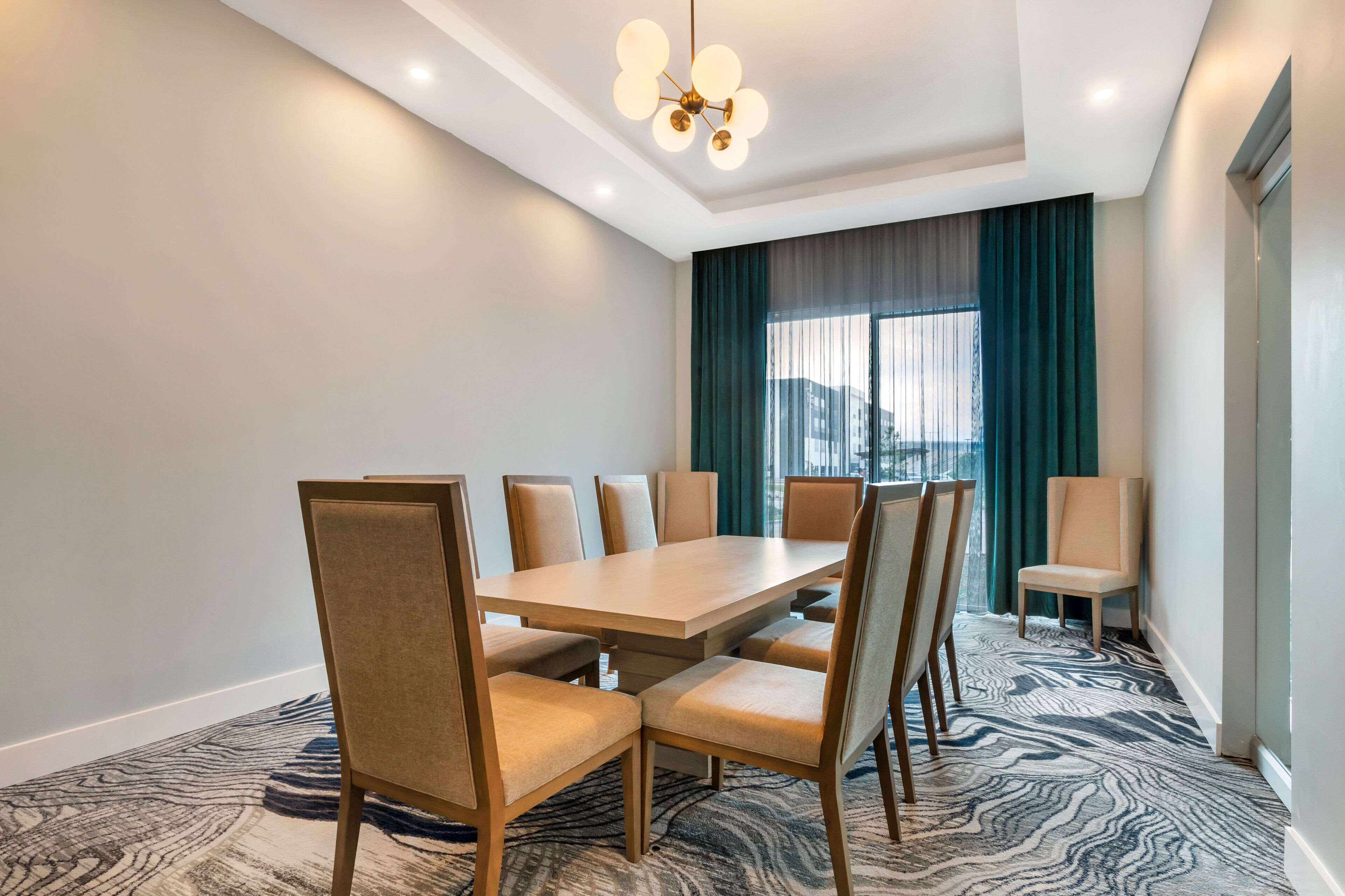 A conference room with a large table and chairs, a patterned carpet, and a view through a window with curtains.