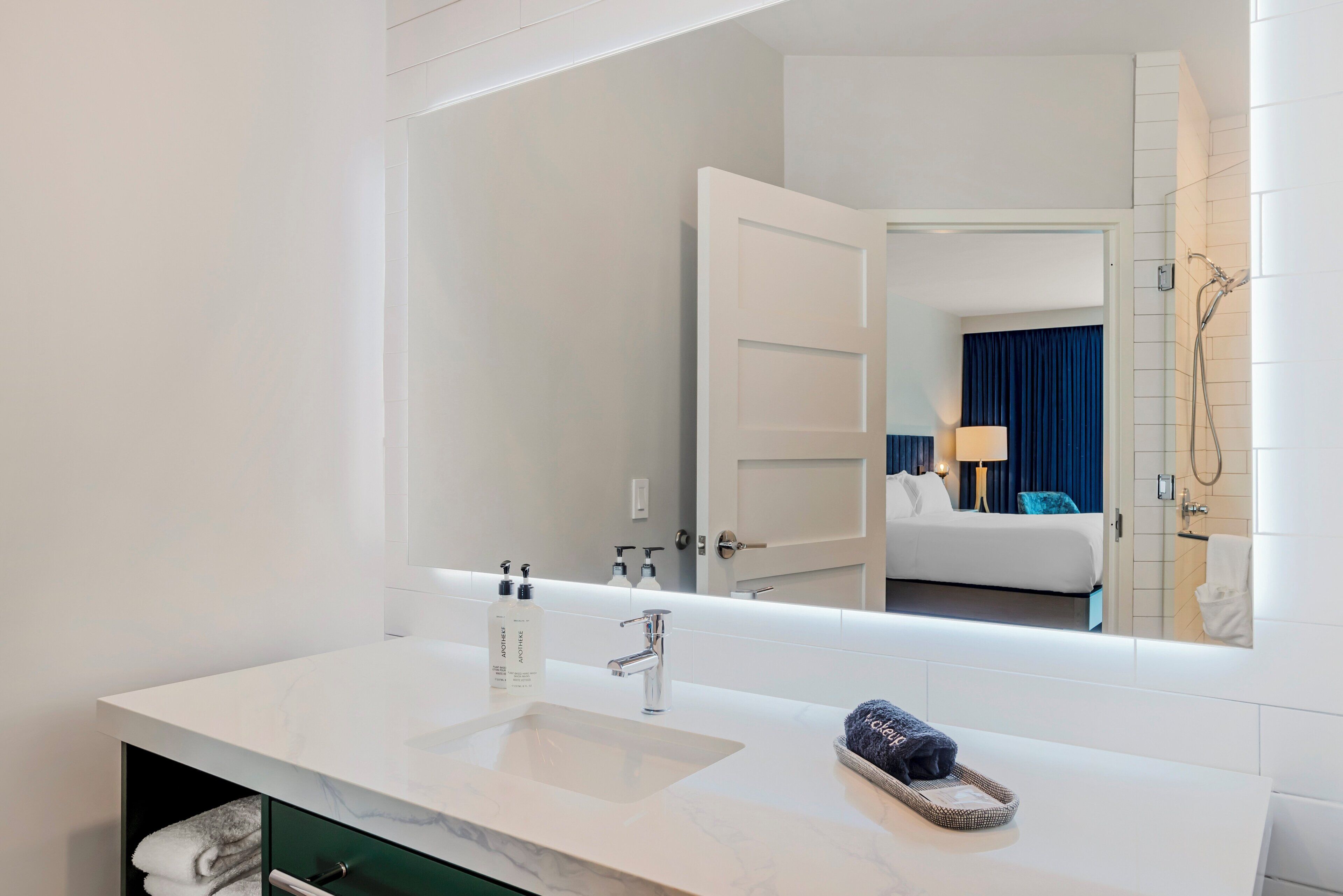 A hotel bathroom with a white sink, a mirror, and a view of a bedroom with a bed and a shower.