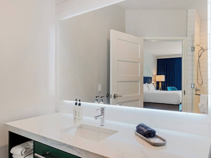 A hotel bathroom with a white sink, a mirror, and a view of a bedroom with a bed and a shower.