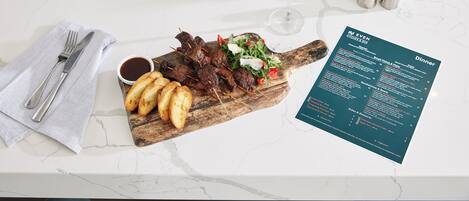 A dining table with a menu, a glass of red wine, skewered meat, roasted potatoes, and a small bowl of sauce.
