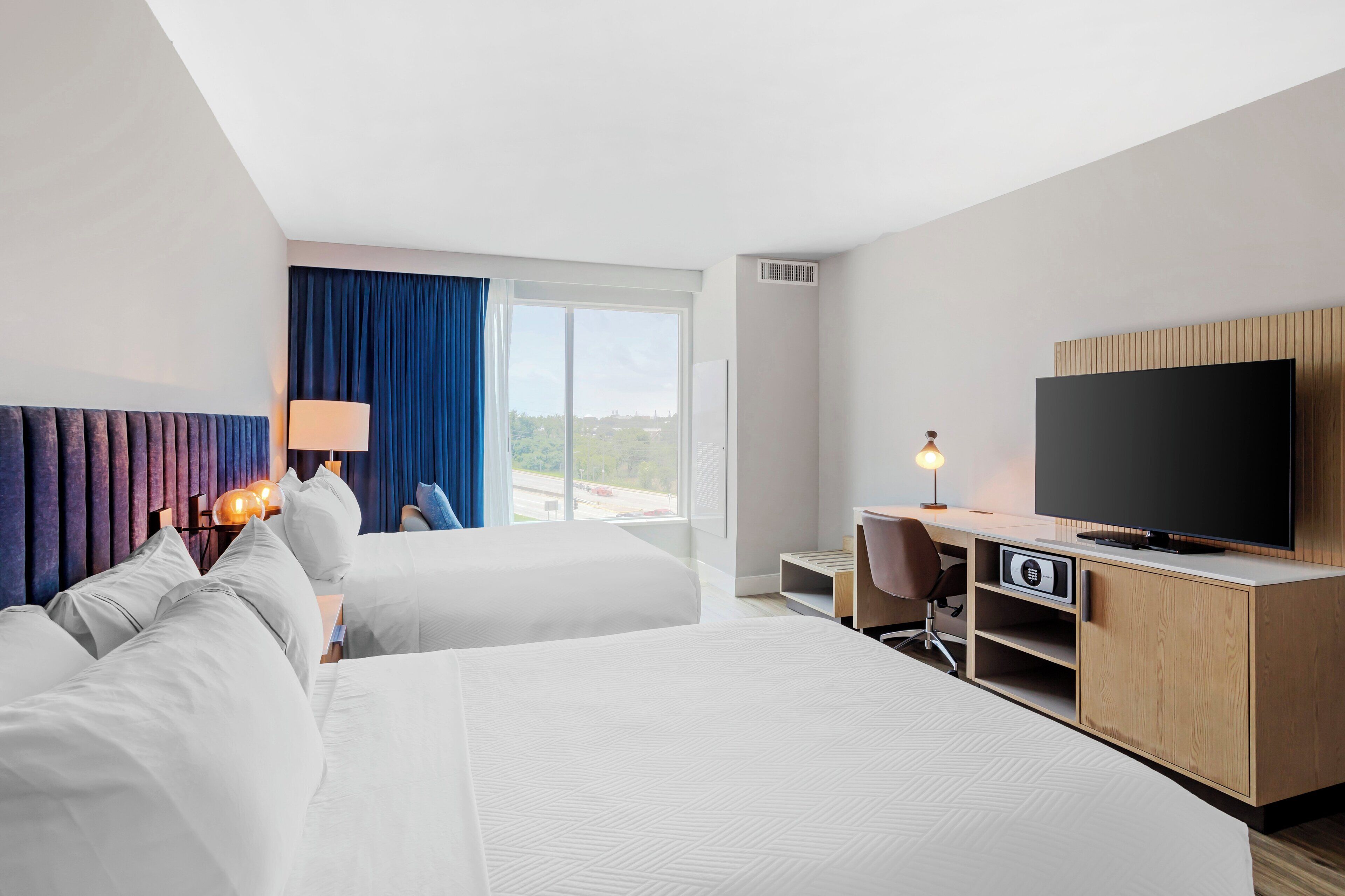 A hotel room with two beds, a desk, a television, and a large window with a view.