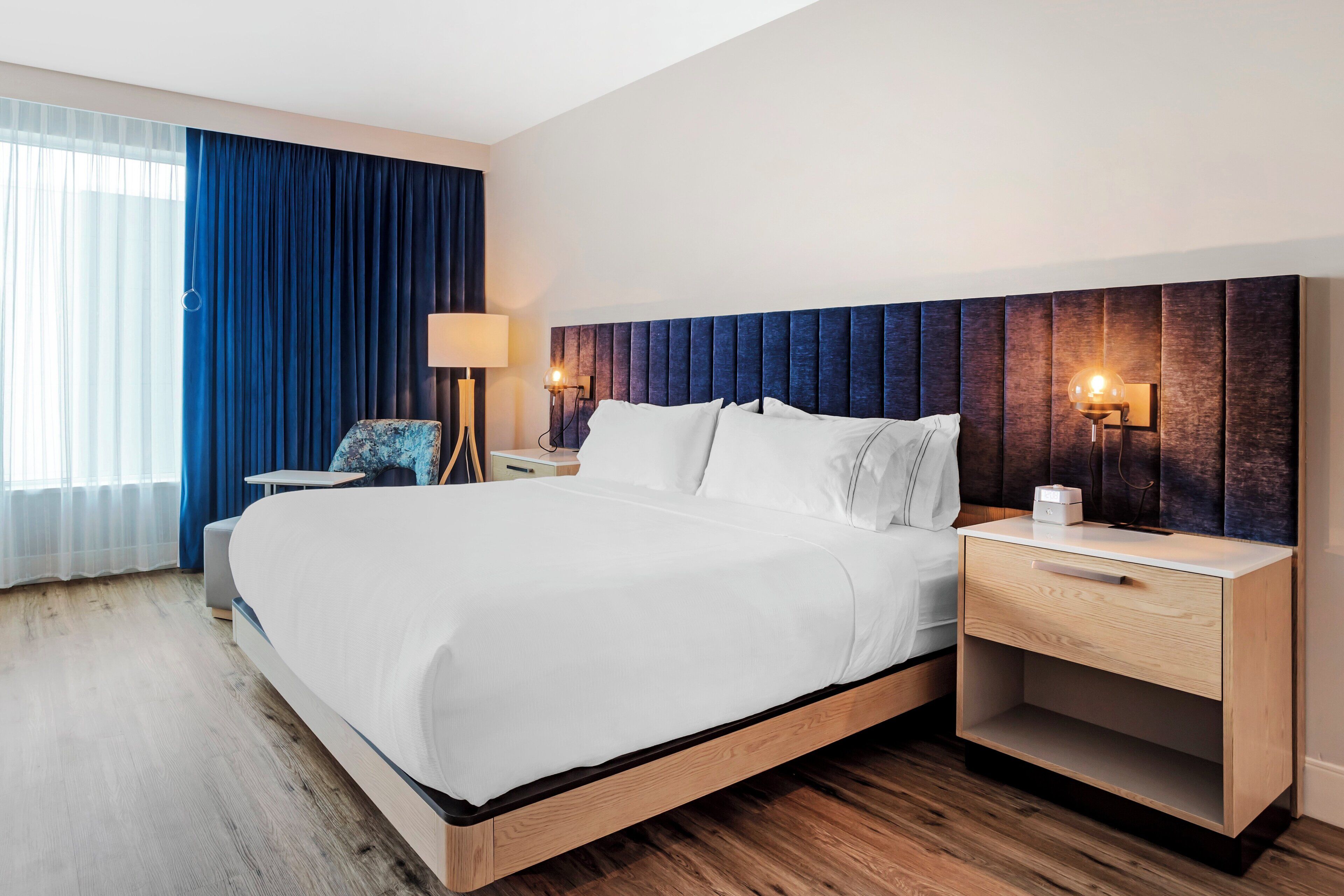 Een moderne hotelkamer met een groot bed, een houten hoofdbord, twee nachtkastjes met lampen, een bijzettafeltje en een raam met blauwe gordijnen.