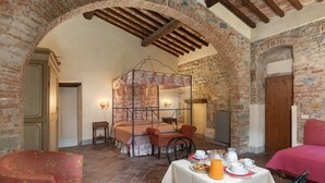 Een rustieke kamer met open bakstenen muren, een houten plafond en een bed met hemelbed. Er staat een tafel gedekt voor het ontbijt, met kopjes, een karaf en een bord eten.