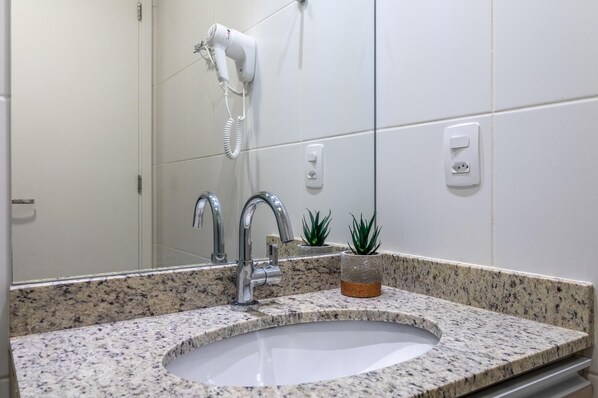 Une salle de bain avec un lavabo doté d’un plan de travail en granit, deux robinets chromés, un sèche-cheveux fixé au mur et deux petites plantes en pot.