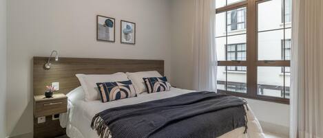 A bedroom with a large bed, a nightstand, a lamp, two framed pictures, and a window with curtains.