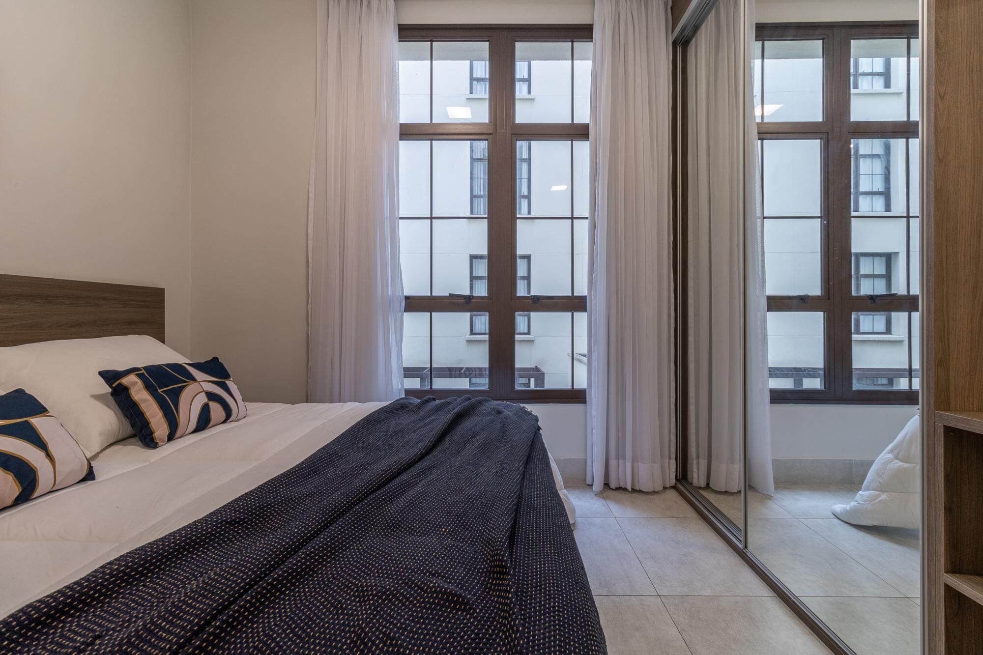 A bedroom with a large window, a bed with a dark patterned blanket, and a wooden headboard.