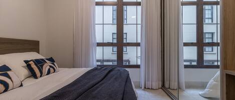 A bedroom with a large window, a bed with a dark patterned blanket, and a wooden headboard.
