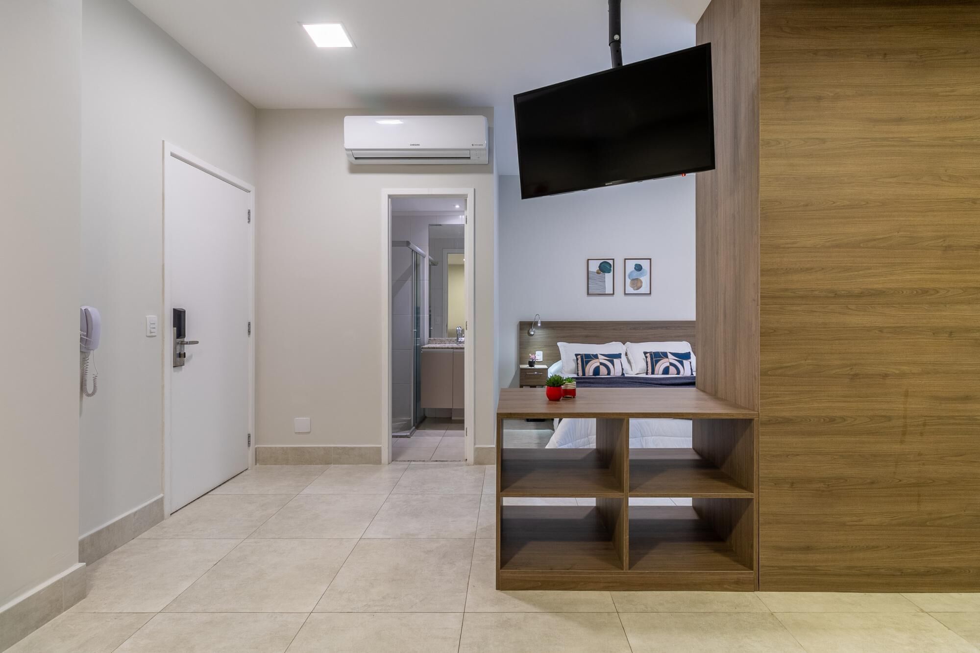 Camera d'albergo con un mobile TV in legno, un letto con cuscini, un tavolino con una pianta e un bagno visibile attraverso una porta aperta.