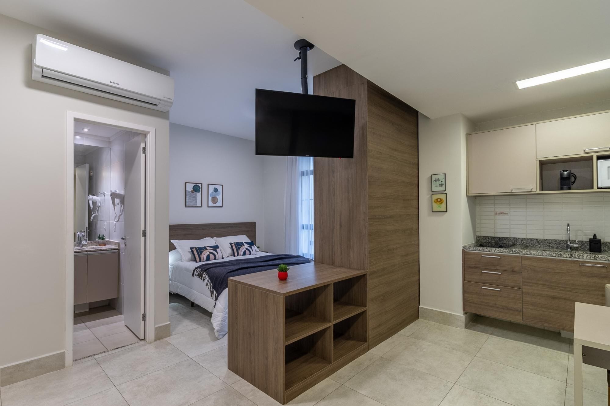 A modern hotel room with a bed, a TV, a small desk, a bathroom, and a kitchenette.