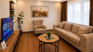 A modern living room with a flat-screen TV, a beige sofa, a round coffee table, a framed picture on the wall, and a potted plant. - Lumiera Suites Hotel (Bursa)