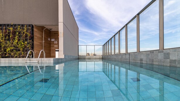 Ein modernes Hallenbad mit klaren blauen Fliesen, einer Holzvertäfelung mit Pflanzen und großen Fenstern mit Blick auf die Skyline der Stadt.