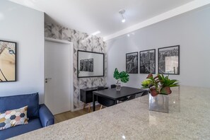 A modern kitchen with a marble countertop, a dining table, and chairs. There are framed pictures on the wall, a potted plant, and a blue sofa in the living area. - Hype104 in S o Paulo (São Paulo)