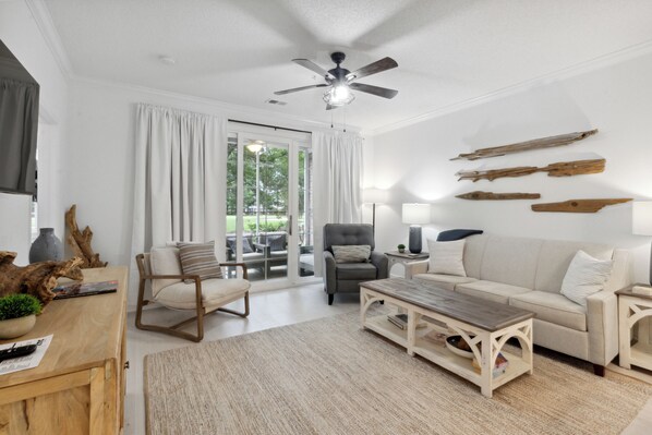 A spacious living room with a sofa, coffee table, and chairs, a ceiling fan, and a view through a glass door to an outdoor area.