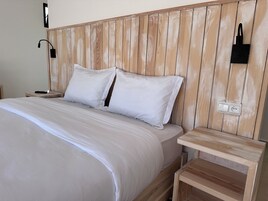 Una cama bien hecha con sábanas blancas, un cabecero de madera, una mesita de noche con una lámpara y un enchufe en la pared.