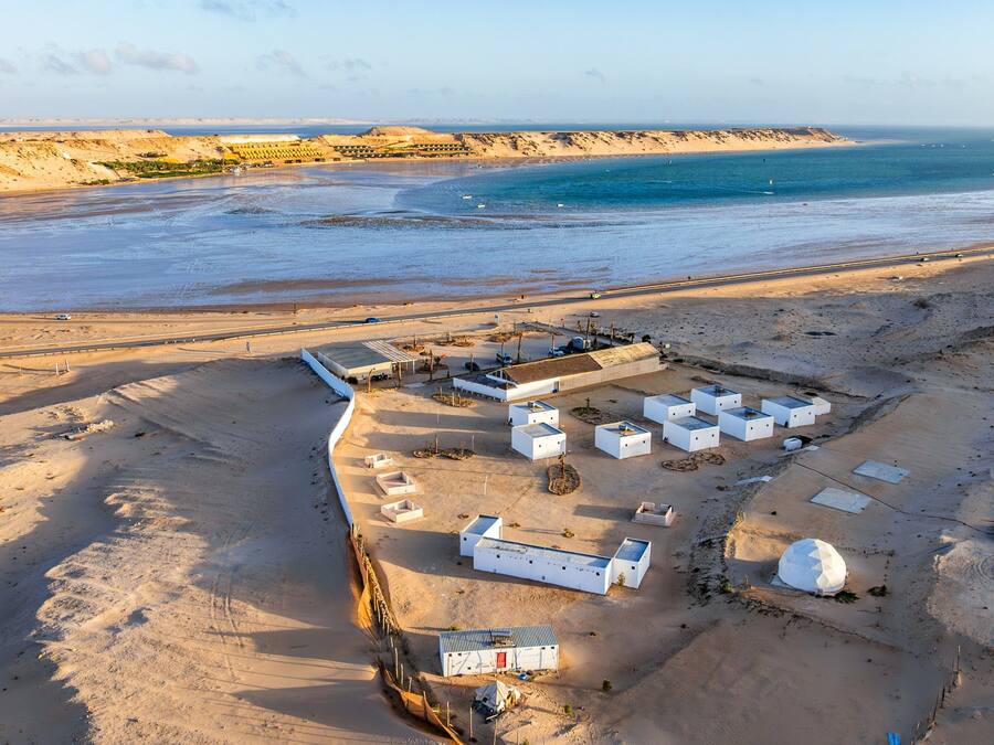 Vista aérea de una zona costera con varios edificios blancos, una playa de arena y una carretera que lleva al mar.