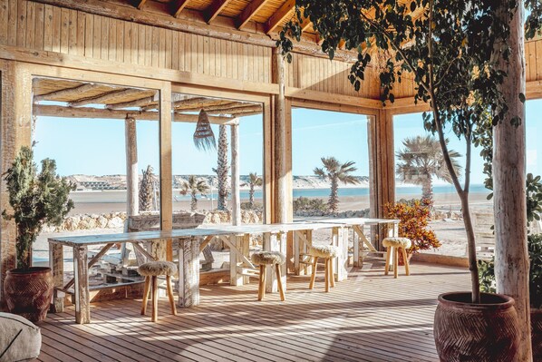 Una terraza de madera con una mesa larga y taburetes, con vista a una playa con palmeras y un cielo despejado.