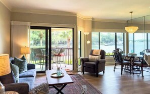 A spacious living area with a sofa, armchair, dining table, and chairs. There is a large window with a view of the outdoors.